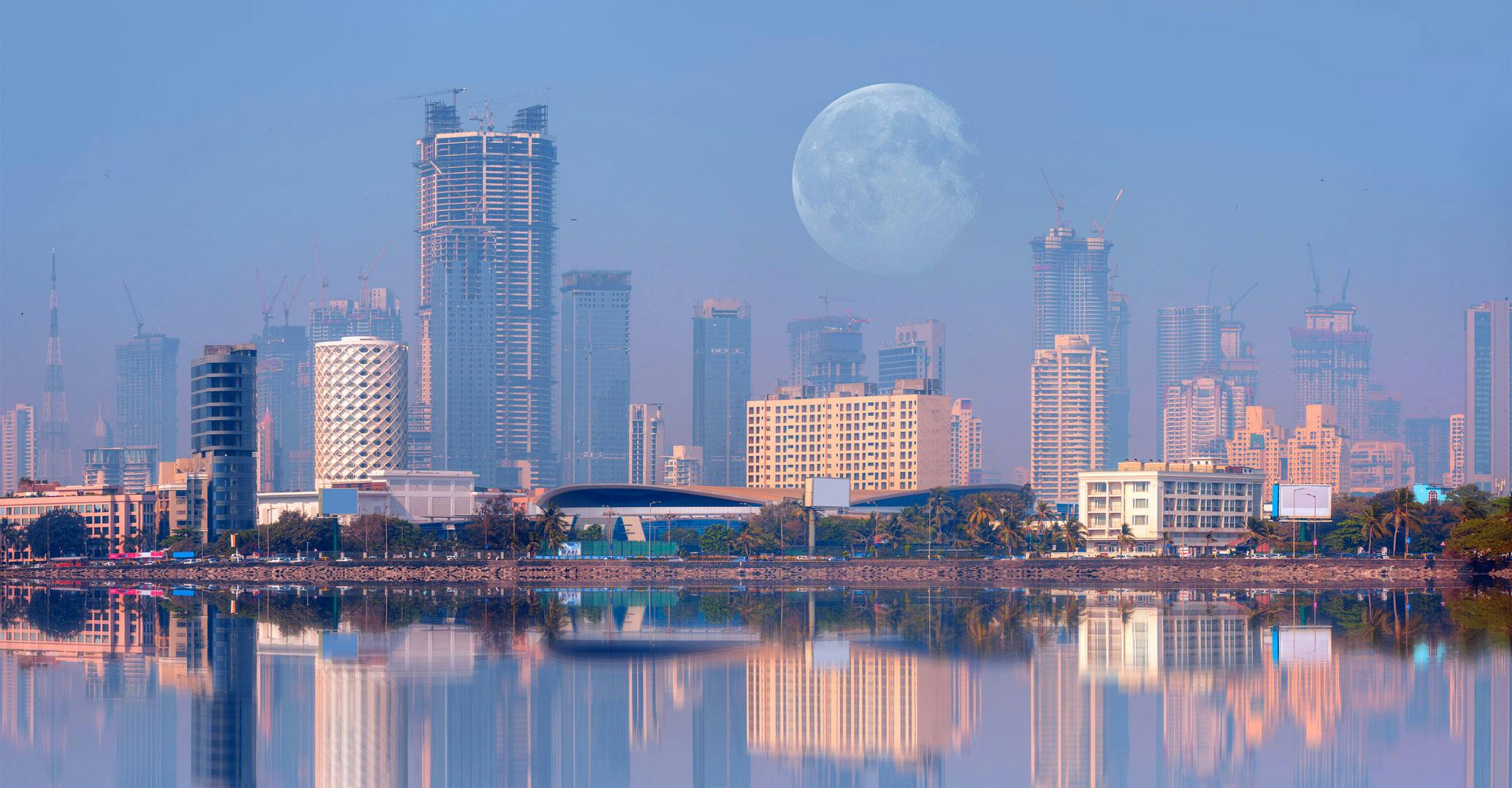 Mumbai cityscape; skyscrapers with cranes on top and the moon in the distance, all reflected on the water