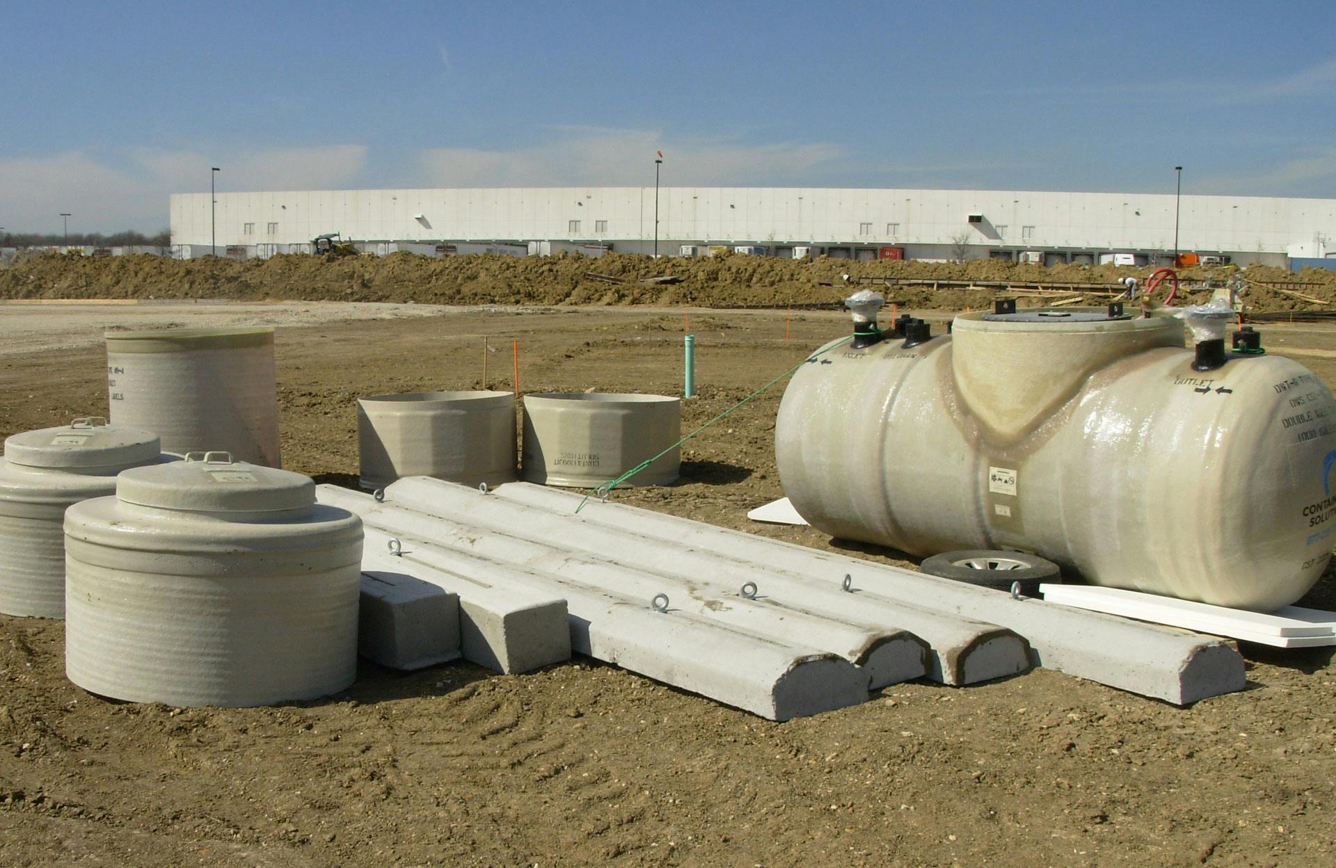 Fiberglass sumps, manways, and deadmen anchors sitting on the ground next to a fiberglass tank at a job site with a building in the background. 